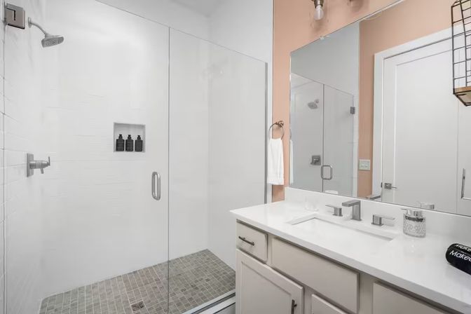 Bright modern bathroom with frameless glass shower, rain showerhead and recessed niche with black bottles, gray tiled shower floor, white vanity with quartz countertop, rectangular mirror and peach accent wall.