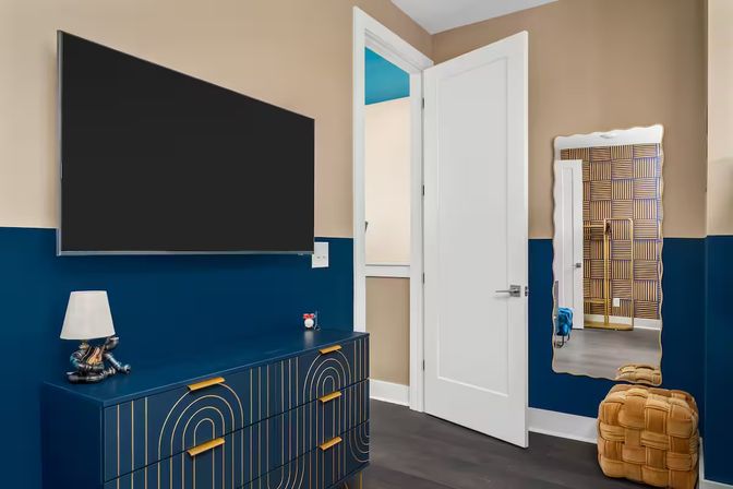 Modern bedroom corner with navy lower walls and beige upper walls, wall-mounted flat-screen TV above a navy dresser with gold geometric handles, white open door, wavy framed mirror and woven tan pouf.