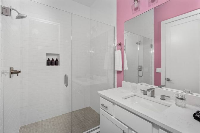 Bright modern bathroom with glass walk-in shower and white subway tile, recessed shampoo niche, pink accent wall, white vanity with quartz countertop and brushed nickel fixtures.