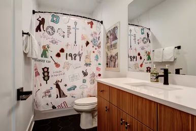 Playful modern apartment bathroom with wood-grain vanity and white countertop, black hex tile floor, white toilet, large mirror, and a colorful collage-style shower curtain with retro icons and numbers.