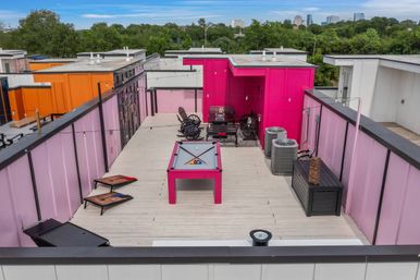 Vibrant magenta urban rooftop deck with billiards table, cornhole boards, lounge seating, air-conditioning units, string lights and a distant city skyline