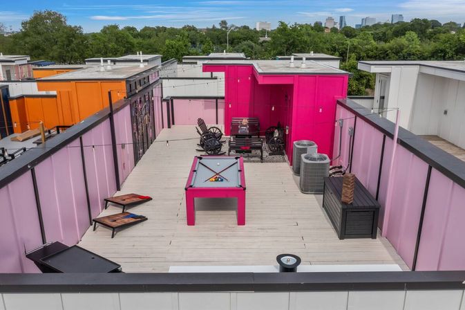 Vibrant magenta urban rooftop deck with billiards table, cornhole boards, lounge seating, air-conditioning units, string lights and a distant city skyline
