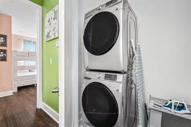 Compact home laundry nook with stacked stainless steel front-load washer and dryer, ironing board and iron, green accent wall and a glimpse of a pastel bedroom with a bunk bed.