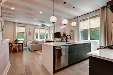 Sunlit open-concept kitchen and living room with white waterfall island and dark green cabinets, three globe pendant lights, stainless dishwasher, cozy sofa, dining nook, hardwood floors, and large windows overlooking trees.