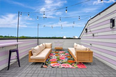 Rooftop patio lounge with wicker sofas facing a vibrant floral rug, pastel lavender-striped siding, hanging string lights and a purple bar stool