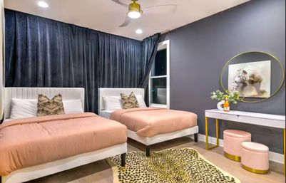 Modern twin bedroom interior with blush-pink duvets on white upholstered beds, dark floor-to-ceiling curtains, gray accent wall with round mirror and tiger art, white vanity with gold legs, pink ottomans, leopard-print rug and ceiling fan.