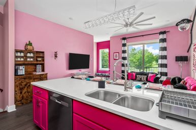 Playful pink open-concept kitchen and living room with hot-pink cabinets, white quartz island and stainless double sink, black-and-white striped curtains, pink sofa, ceiling fan and balcony view of green trees.