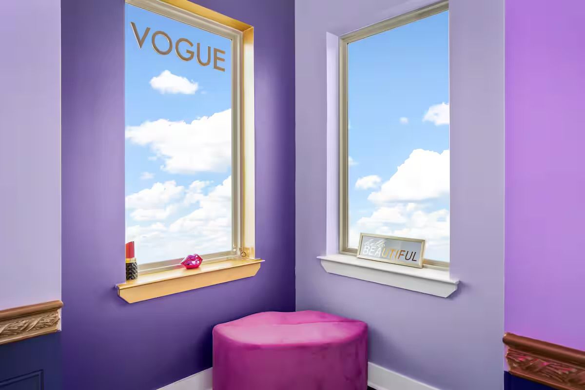 Bright purple vanity nook with two tall windows showing blue sky and clouds, gold 'VOGUE' window decal, lipstick and lip-shaped decor on the sill, a framed 'BEAUTIFUL' sign, and a pink round ottoman.
