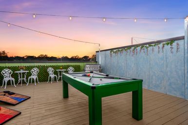 Sunset rooftop patio with a bright green pool table, twinkling string lights, white bistro chairs and tables, cornhole boards, a stainless grill and decorative ivy on a wooden deck.
