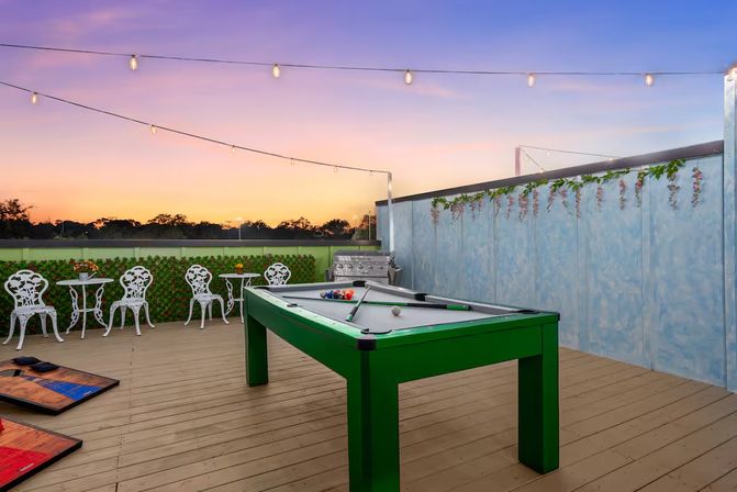 Sunset rooftop patio with a bright green pool table, twinkling string lights, white bistro chairs and tables, cornhole boards, a stainless grill and decorative ivy on a wooden deck.