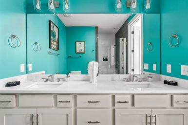 Bright turquoise master bathroom with white double-sink vanity, quartz countertop, chrome faucets and hardware, large mirror, sculptural head decor, framed art and glass-enclosed shower reflected in mirror.