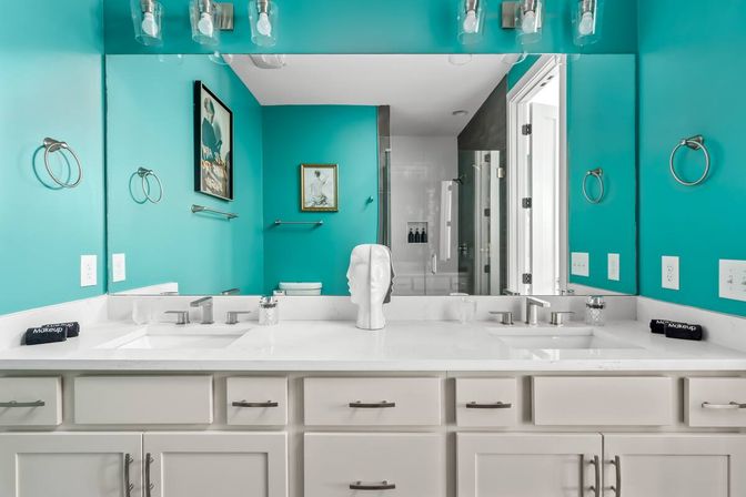 Bright turquoise master bathroom with white double-sink vanity, quartz countertop, chrome faucets and hardware, large mirror, sculptural head decor, framed art and glass-enclosed shower reflected in mirror.