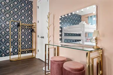 Glam dressing nook with lighted vanity mirror, glass-top gold console, pink velvet stools, brass coat rack, dark floral wallpaper and reflected white bunk beds.