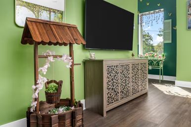 Sunlit living room corner with a wooden decorative wishing-well planter draped in pink blossoms, a carved light-wood console with filigree doors under a wall-mounted TV, green-painted walls, window and floral accents.