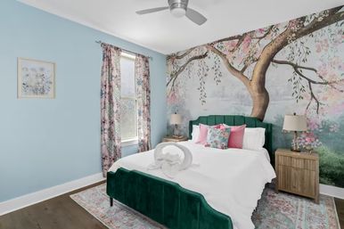 Bright cozy bedroom with green velvet bed and white bedding, towel swan on the bed, large tree and cherry blossom mural across the wall, light blue walls, floral curtains, pink accent pillows, wooden nightstands and ceiling fan