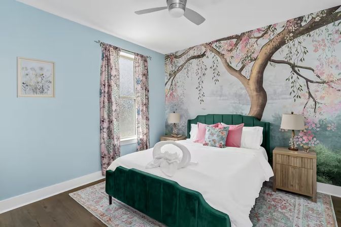 Bright cozy bedroom with green velvet bed and white bedding, towel swan on the bed, large tree and cherry blossom mural across the wall, light blue walls, floral curtains, pink accent pillows, wooden nightstands and ceiling fan