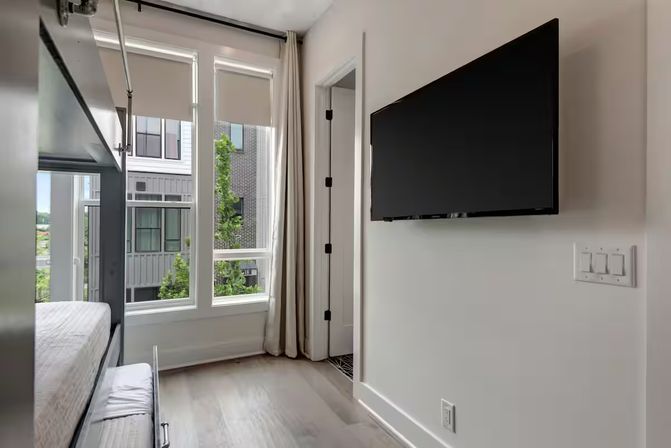 Cozy modern apartment bedroom with a bunk bed, large floor-to-ceiling windows showing neighboring buildings and trees, wall-mounted flat-screen TV and light hardwood floors.