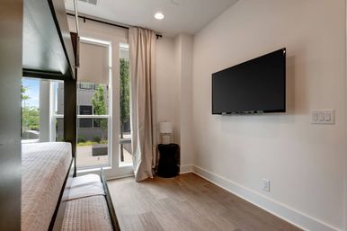 Bright modern apartment bedroom with built-in bunk bed, wall-mounted flat-screen TV, floor-to-ceiling windows with roller shades and curtains, small black side table with lamp, and wood-look flooring.