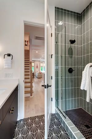 Modern home bathroom with glass walk-in shower clad in vertical green tiles, black hexagon shower floor, geometric black-and-white floor tiles, white vanity and open door showing staircase and living room beyond.