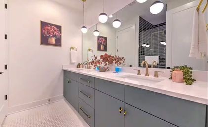 Modern residential bathroom with teal-gray double-sink vanity, white quartz countertop, brass faucets, round pendant lights, large mirror, hexagon tile floor, and floral decor