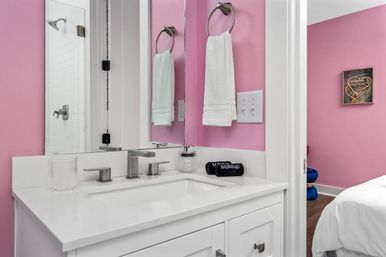 Playful pink bathroom with white vanity and quartz countertop, modern brushed-nickel faucet over rectangular sink, round towel ring holding a white towel, mirrored wall and glass shower door, small makeup pouch on the counter and a bedroom visible through the doorway.