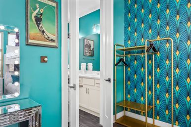 Vibrant dressing area with turquoise walls, art-deco blue-and-gold fan-pattern wallpaper, gold open clothing rack, lighted vanity mirror, framed art, and white double doors opening to a bathroom vanity.