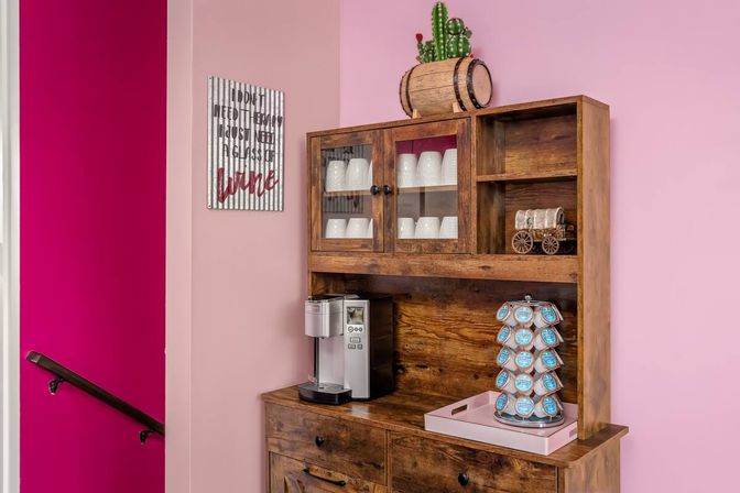 Cheerful pink-walled coffee nook with a rustic wooden hutch, silver single-serve coffee maker, rotating K-Cup carousel, glass-doored cabinet of white mugs and barrel cactus decor.