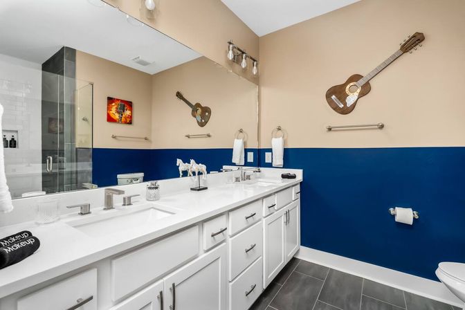 Bright modern master bathroom with white double-sink vanity and quartz countertop, navy blue lower walls and beige upper walls, glass walk-in shower, acoustic guitar wall decor and gray floor tiles — stylish residential bathroom interior.