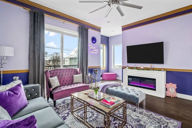 Sunlit modern glam living room in lavender and purple tones featuring a tufted magenta loveseat, gray sofa with purple pillows, patterned area rug, glass-top coffee table, wall-mounted TV above an electric fireplace with purple flame, large window with gray curtains and ceiling fan.