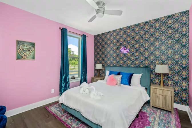 Colorful modern eclectic bedroom with pink walls and a patterned floral accent wall, teal upholstered headboard, white bed linens with blue and pink throw pillows, teal curtains, wooden nightstands, neon script sign, ceiling fan, hardwood floor and vibrant area rug.