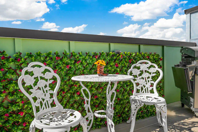 Sunlit rooftop balcony with white ornate cast-iron bistro table and chairs, boot vase of yellow flowers, green privacy hedge with red blooms and a compact gas grill under a bright blue sky.