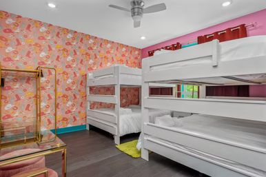 Playful kids' bedroom with two white double bunk beds, vibrant pink patterned wallpaper, dark hardwood floors, ceiling fan, red curtains, and a gold clothing rack