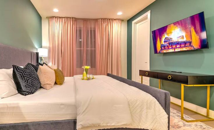 Cozy modern bedroom with a plush gray bed and white layered bedding, velvet and metallic throw pillows, emerald green accent wall, blush pink floor-length curtains, wall-mounted TV displaying a fireplace, and a black console table with gold legs.