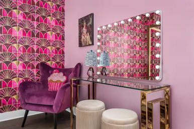 Pink glam dressing nook with art-deco gold and fuchsia wallpaper, illuminated Hollywood vanity mirror, glass-top gold console table, velvet plum armchair with cupcake pillow, and two round cream ottomans.