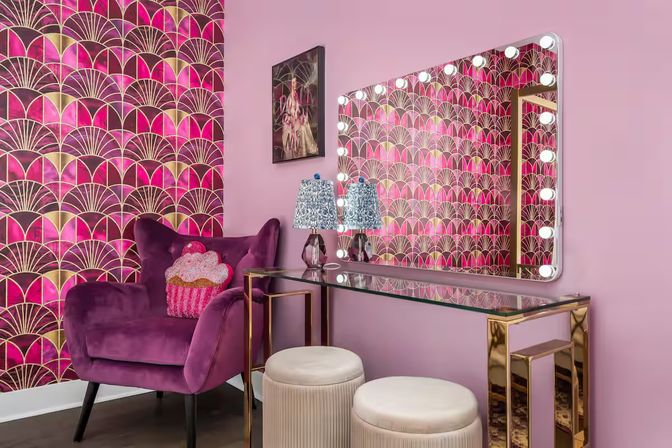 Pink glam dressing nook with art-deco gold and fuchsia wallpaper, illuminated Hollywood vanity mirror, glass-top gold console table, velvet plum armchair with cupcake pillow, and two round cream ottomans.