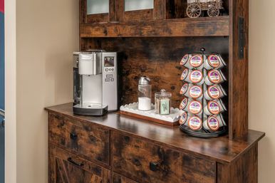 Coffee station on a rustic wooden cabinet: silver single-serve coffee maker, rotating rack of single‑serve coffee pods, glass sugar jar and creamer on a decorative tray, countertop setup for morning brews.