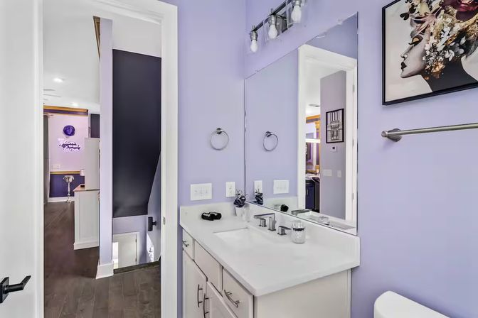 Lavender bathroom interior with white vanity and quartz countertop, chrome faucet and fixtures, large mirror, floral-profile wall art, towel bar, and open doorway leading to dark hardwood hallway and stairs.