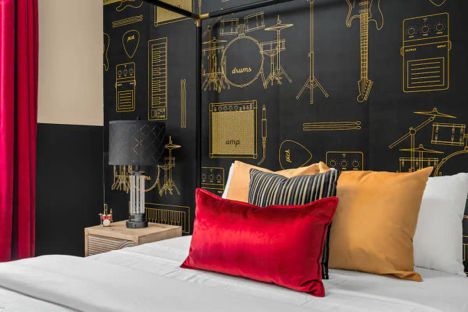 Boutique music-themed hotel room with black-and-gold instrument mural, red velvet accent pillow, striped and mustard throw pillows, and a bedside lamp on a wooden nightstand.