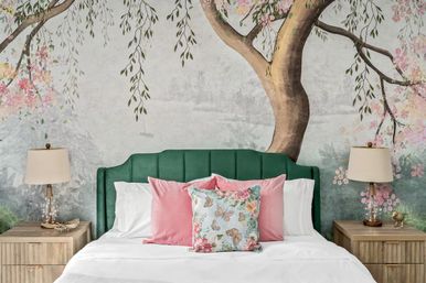 Cozy bedroom interior with emerald green upholstered headboard, large floral tree wall mural, crisp white bedding and pink plus butterfly-print accent pillows, wooden nightstands and glass table lamps.