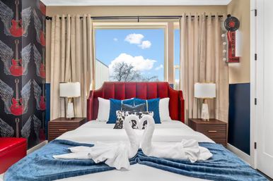 Rock-themed boutique hotel bedroom with red upholstered headboard, guitar-and-wing wallpaper, swan towel art on a white bed layered with a blue throw, wooden nightstands and a window showing a sunny sky view.