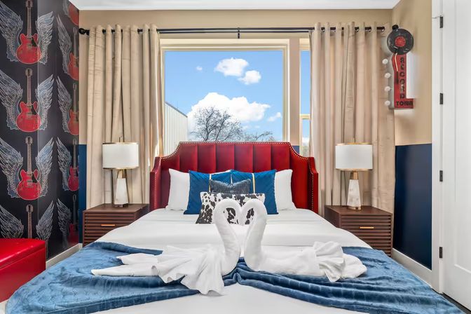 Rock-themed boutique hotel bedroom with red upholstered headboard, guitar-and-wing wallpaper, swan towel art on a white bed layered with a blue throw, wooden nightstands and a window showing a sunny sky view.