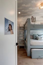 Coastal-inspired kids' bedroom with gray built-in bunk beds, ladder and trundle drawer, cloud-pattern wallpaper and ceiling, hardwood floors, woven pendant light, and a smiling woman poster on the door.