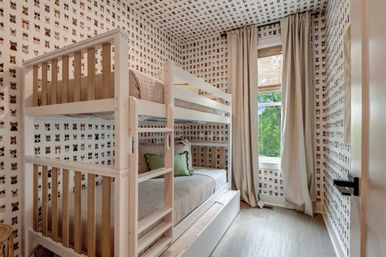 Cozy kids' bedroom with white wooden bunk beds and trundle, gray bedding with a green accent pillow, playful cat-face wallpaper on walls and ceiling, and a tall window with beige curtains showing leafy outdoor view.