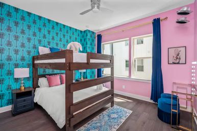 Vibrant kids' bedroom with dark wood bunk bed, teal guitar-patterned wallpaper, pink walls, navy curtains, blue velvet stool, vanity mirror lights and hardwood floors lit by natural window light.