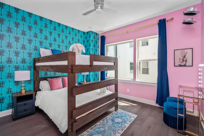 Vibrant kids' bedroom with dark wood bunk bed, teal guitar-patterned wallpaper, pink walls, navy curtains, blue velvet stool, vanity mirror lights and hardwood floors lit by natural window light.