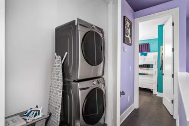 Stacked metallic washer and dryer in a compact laundry closet with ironing board and laundry basket, purple hallway opening to a teal-accented bedroom with a white bunk bed.