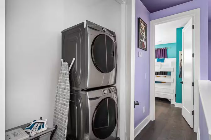 Stacked metallic washer and dryer in a compact laundry closet with ironing board and laundry basket, purple hallway opening to a teal-accented bedroom with a white bunk bed.