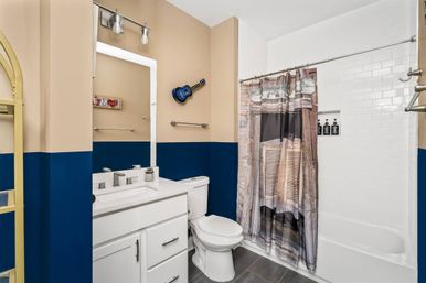Residential modern bathroom interior with two-tone navy and beige walls, white vanity and sink, white toilet, subway-tiled bathtub with printed shower curtain, gray floor tiles and a small blue wall-mounted ukulele.