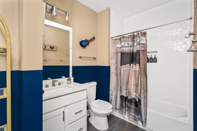 Residential modern bathroom interior with two-tone navy and beige walls, white vanity and sink, white toilet, subway-tiled bathtub with printed shower curtain, gray floor tiles and a small blue wall-mounted ukulele.
