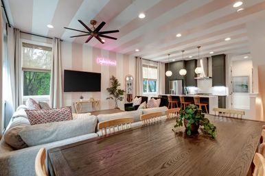 Bright open-concept living room and kitchen with striped ceiling, large wood dining table and plant centerpiece, gray sectional sofa, wall-mounted TV, a pink neon wall sign, pendant lights over a kitchen island with bar stools, and windows showing trees outside.